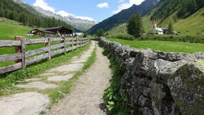 Sentiero di montagna che conduce a una baita di legno con un muro in pietra su un lato e una chiesa con tetto rosso in lontananza.