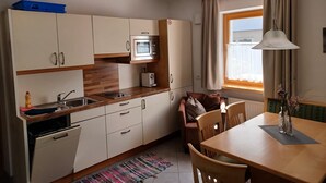 Cocina compacta con microondas, pileta y muebles de madera. Hay una mesa de comedor con sillas y un ventanal con cortinas.