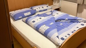 Una cama individual con colcha azul y blanca estampada, cabecera de madera, mesita de noche, ventana con cortinas y un radiador.