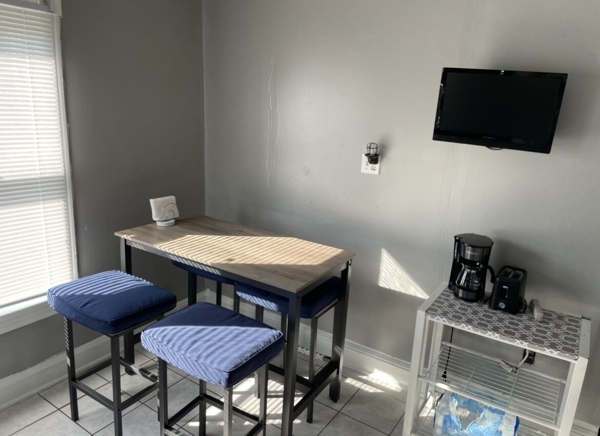 A small, well-lit room with a table and two stools, a mounted television, and a coffee maker on a shelf.