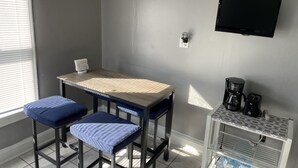 Una habitación pequeña y bien iluminada con una mesa, dos taburetes, un televisor montado y una cafetera en un estante.