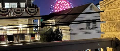 A balcony with a railing, overlooking a street with parked cars and a building with festive lights.