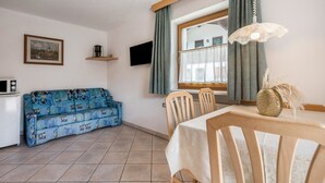 Ein Wohnzimmer mit einer Couch, einem Esstisch mit Stühlen, einem Fernseher und einem Fenster mit Vorhängen.