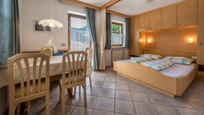 Ein Schlafzimmer mit Bett, Holz-Kopfteil, Nachttisch, Stuhl, Fenster mit Vorhängen und Blick ins Freie.