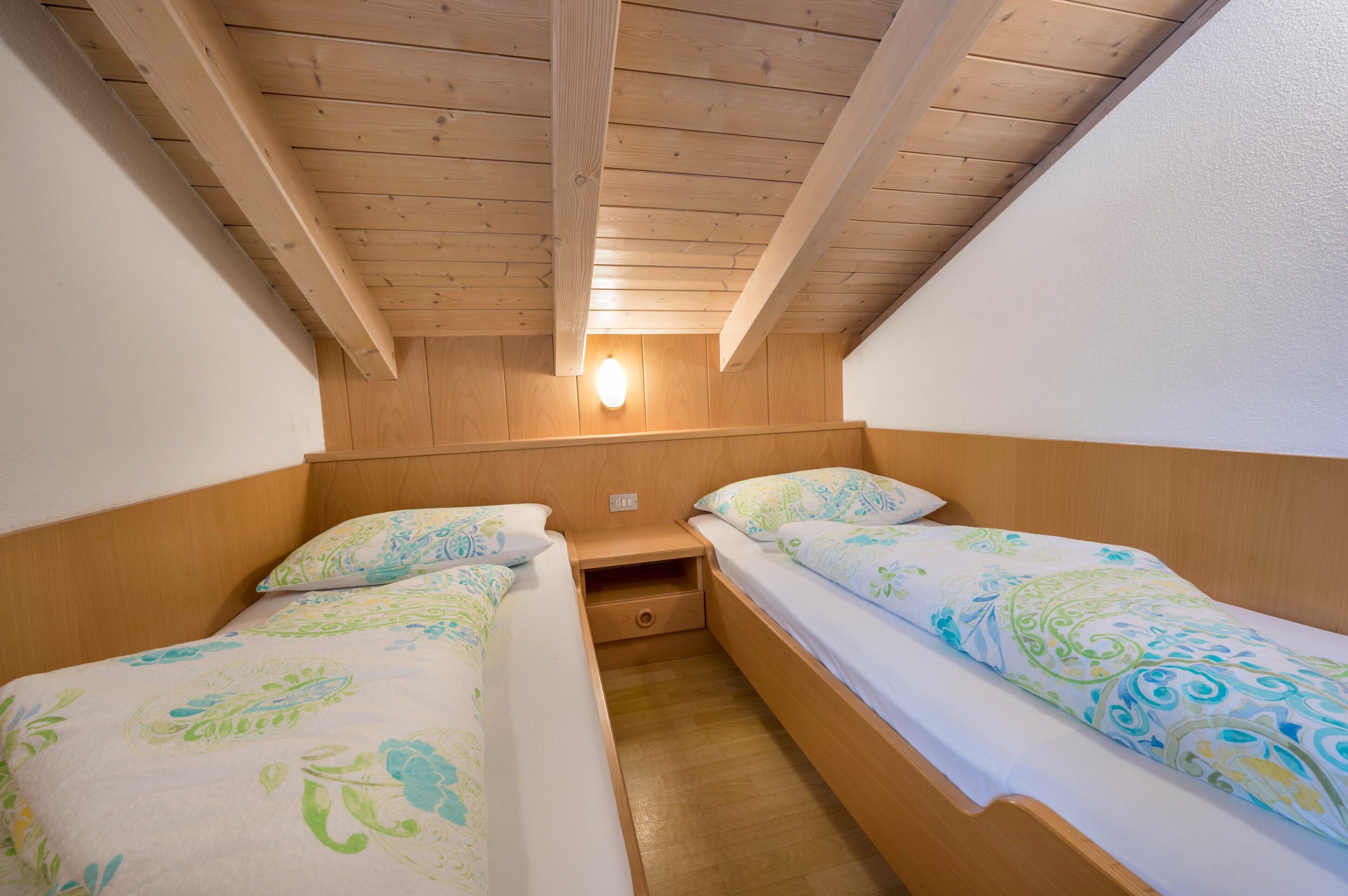 Ein kleiner, gemütlicher Raum mit zwei Einzelbetten, Holzbalkendecke und einer an der Wand befestigten Lampe.
