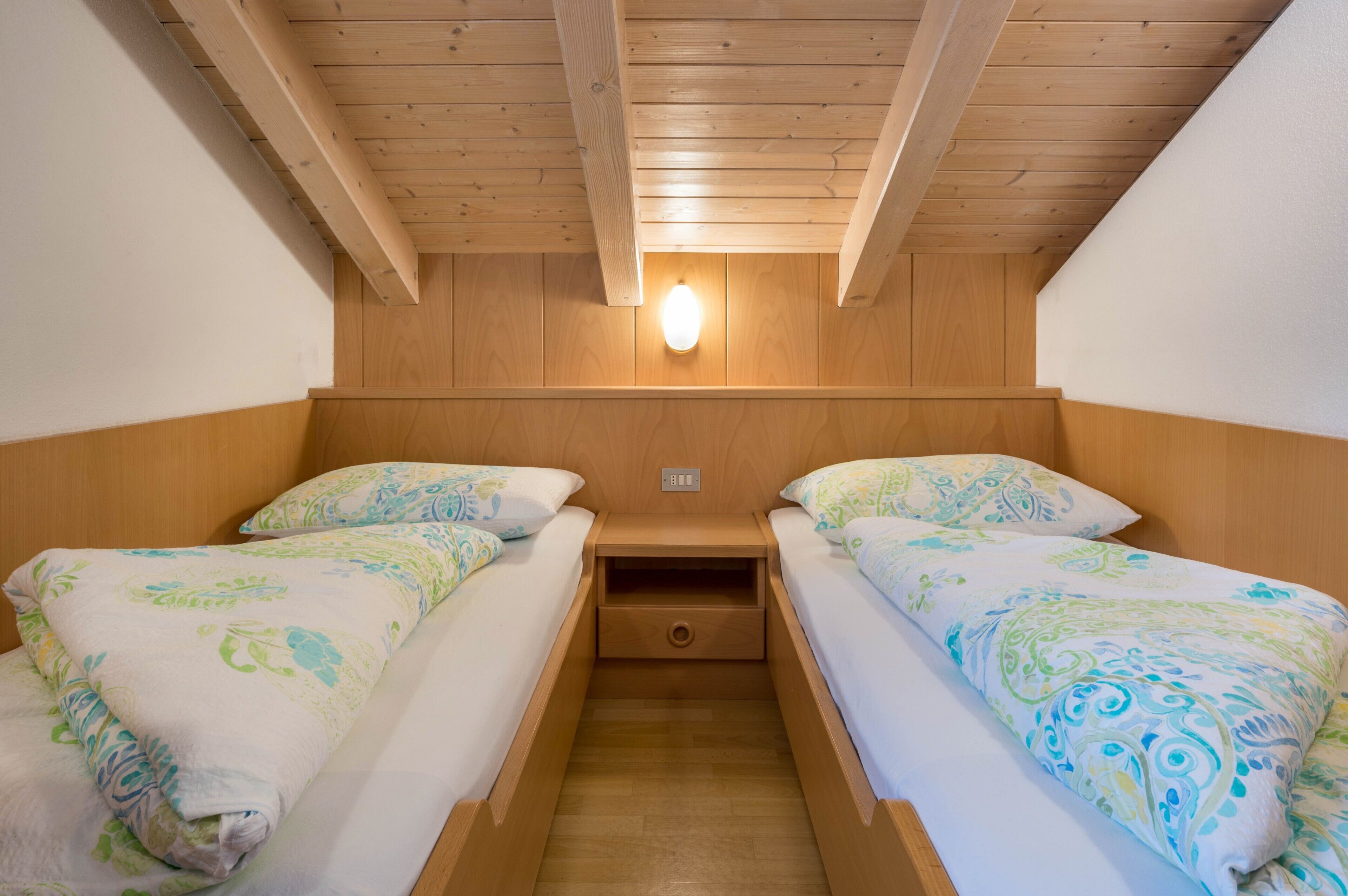 Een kamer met twee bedden, houten balken in het plafond, een wandlamp en een klein lade onder de bedden.