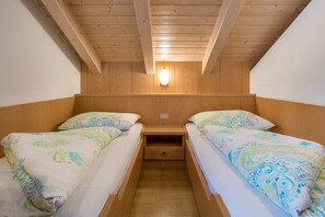 Quarto com duas camas, vigas de madeira no teto, uma luminária na parede e uma gaveta pequena sob as camas.