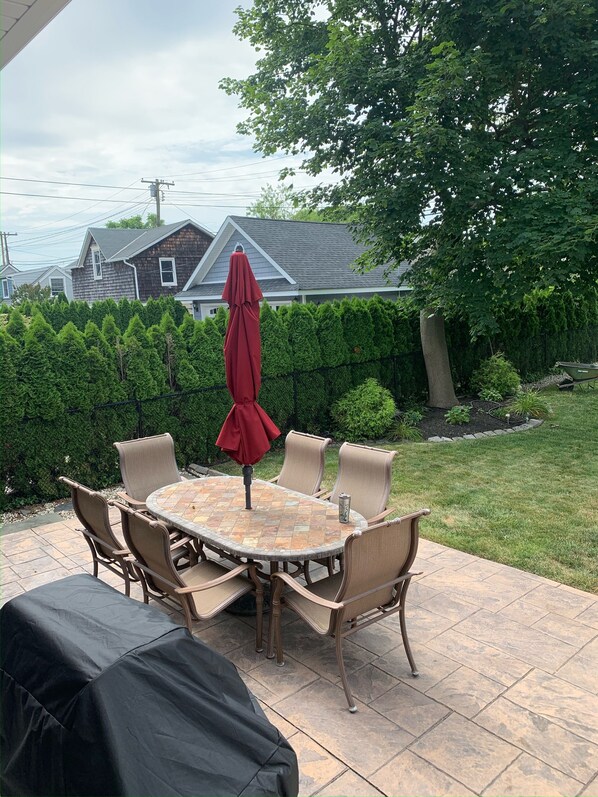 An outdoor patio with a round table and chairs, a covered grill, and a view of a house and trees in the background. - Belmar House 2 blocks from beach (Belmar)