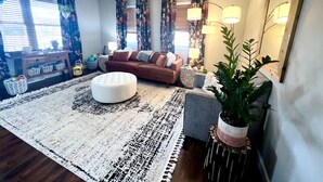 Una sala de estar con sofá, reposapiés y un tapete grande. Hay dos lámparas, un espejo y una planta en maceta.