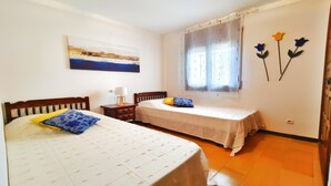 Un dormitorio con dos camas, una mesita de noche, una ventana con cortinas traslúcidas y un adorno floral en la pared.