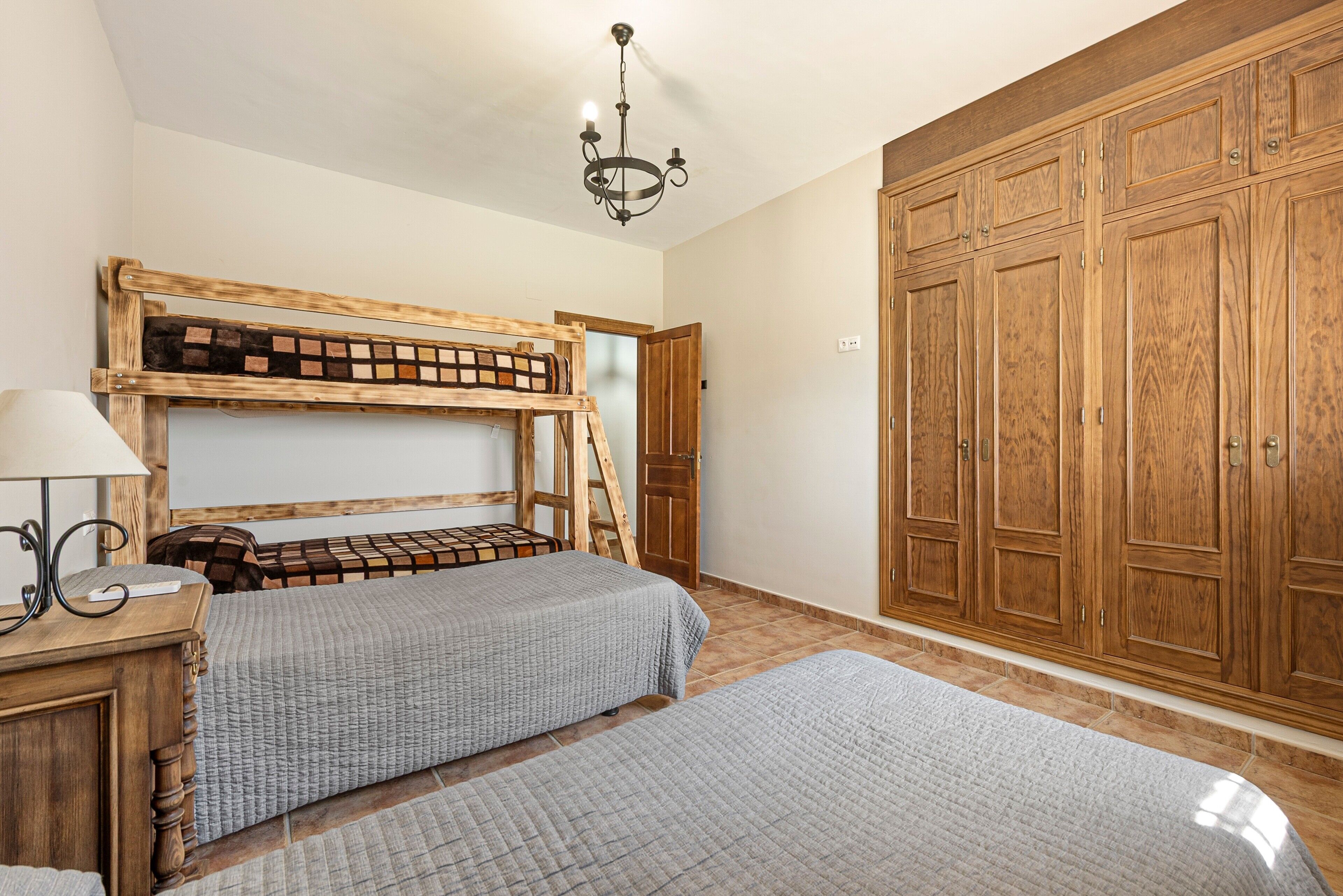 Une chambre avec des lits superposés, une armoire en bois, un lustre et une table de chevet avec une lampe.