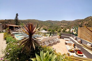 A pool area with lounge chairs, a dining table with food, and a mountainous backdrop.