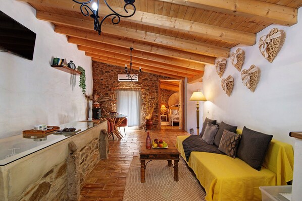 A cozy room with a stone wall, wooden ceiling beams, a sofa, a table with fruit, and a TV.