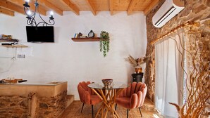 A rustic interior with a stone wall, wooden ceiling beams, a round glass table with chairs, a mounted TV, and a potted plant. - Country House 'Quinta Do Silêncio T2' with Mountain View, Wi-Fi and Air Conditioning (Tavira)