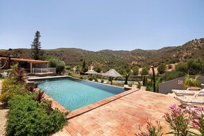A swimming pool with a patio area, surrounded by greenery and hills in the background.