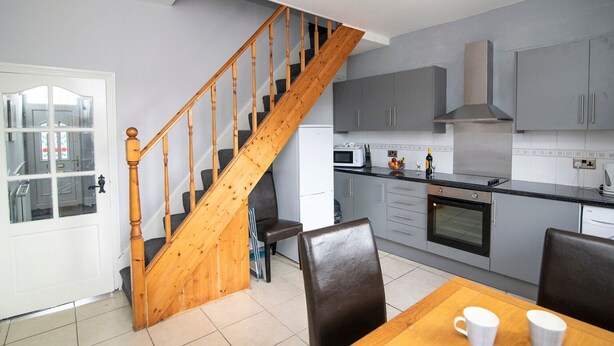 A kitchen with grey cabinets, a wooden staircase, a dining table with two chairs, and a microwave. - Bolton 2 Bedroom House | Free Parking & WiFi | Ideal for Long Stays (Tonge Fold)