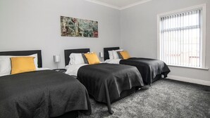 A hotel room with three beds, a large window with blinds, and a cityscape painting on the wall. - Bolton 2 Bedroom House | Free Parking & WiFi | Ideal for Long Stays (Tonge Fold)