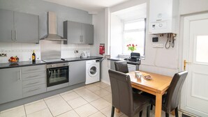 A modern kitchen with grey cabinets, a built-in oven, a washing machine, and a dining area with a wooden table and chairs. - Bolton 2 Bedroom House | Free Parking & WiFi | Ideal for Long Stays (Tonge Fold)