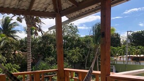 A hammock under a wooden pergola with a tiled floor, surrounded by greenery and a clear sky.