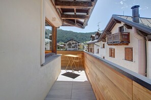 A narrow corridor with a wooden bench and a table, leading to a balcony with a view of a village and mountains.