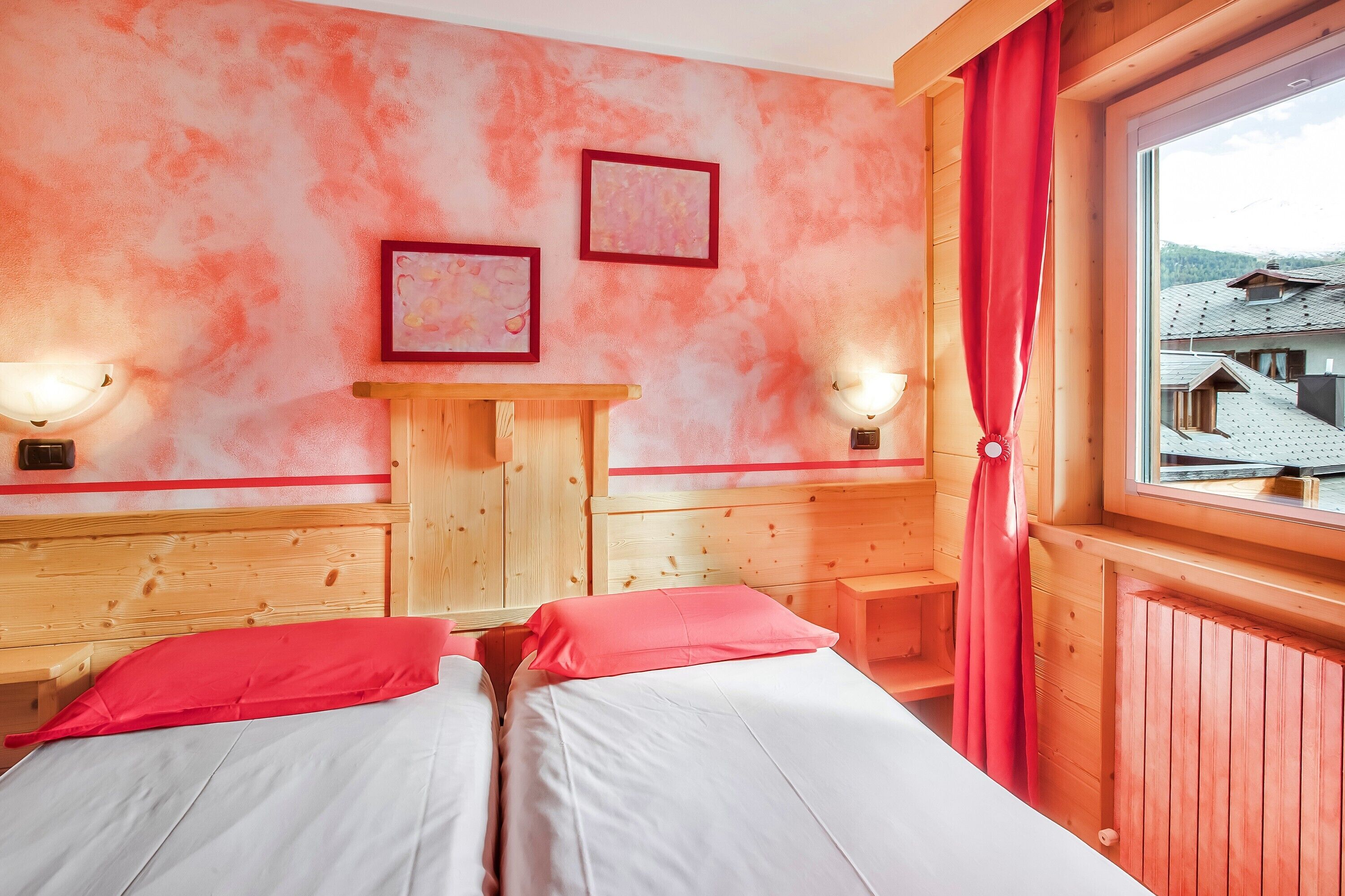 A room with two beds, wooden headboards, a wooden desk, two wall-mounted lamps, and a window with a view of buildings.