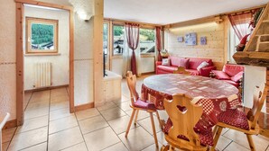 Ein gemütliches Zimmer mit Holzvertäfelung, einem runden Tisch mit kariertem Tischläufer, einer roten Couch und Blick auf einen Wald durch ein Fenster.