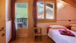 Een houten kamer met een bed, een klein tafeltje, een raam met gordijnen en uitzicht op een houten gebouw en bomen.