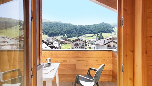 Een balkon met houten vloer, een tafel en een stoel, met uitzicht op een dorp en beboste heuvels.