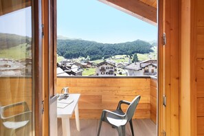A balcony with a wooden floor, a table, and a chair, overlooking a village and forested hills.