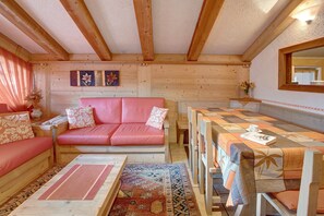 Une pièce chaleureuse avec un plafond en bois, un canapé rouge, une table à manger recouverte d’une nappe à motifs et un miroir au mur.