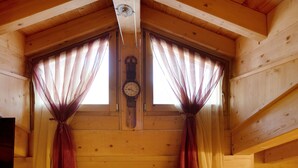 Ein Holzblockbau-Innenraum mit einer Uhr an der Wand, zwei Fenstern mit Vorhängen und einer Tür.