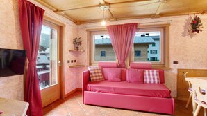 Ett mysigt rum med en rosa soffa, en tv, ett fönster med gardiner och ett träpanelat tak.