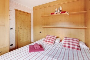 A wooden bedroom with a bed, two pillows, a rolled towel, and a shelf with decorative items.