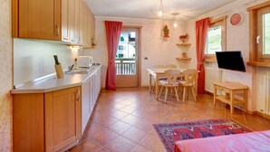 Cocina compacta con armarios de madera, zona de comedor con mesa y sillas, televisor y vista a través de una puerta.