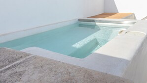 Een klein, rechthoekig zwembad met helder water, omringd door witte muren en een houten terras.