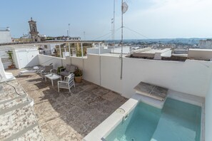 A rooftop terrace with a swimming pool, lounge chairs, and a view of a cityscape including a church tower.