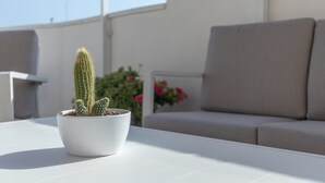 Een cactus in een witte pot op een witte tafel, een beige bank en een witte muur met een plant op de achtergrond.