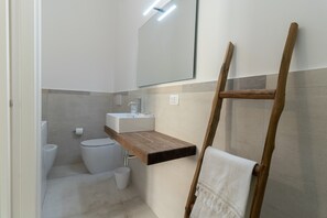 A bathroom with a toilet, sink, mirror, and wooden shelf.