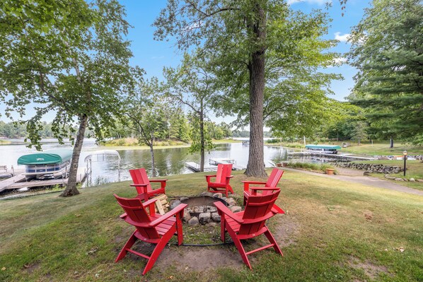 Zona de estar junto al lago con sillas Adirondack rojas alrededor de un brasero, con vista a un lago tranquilo con botes y un muelle.