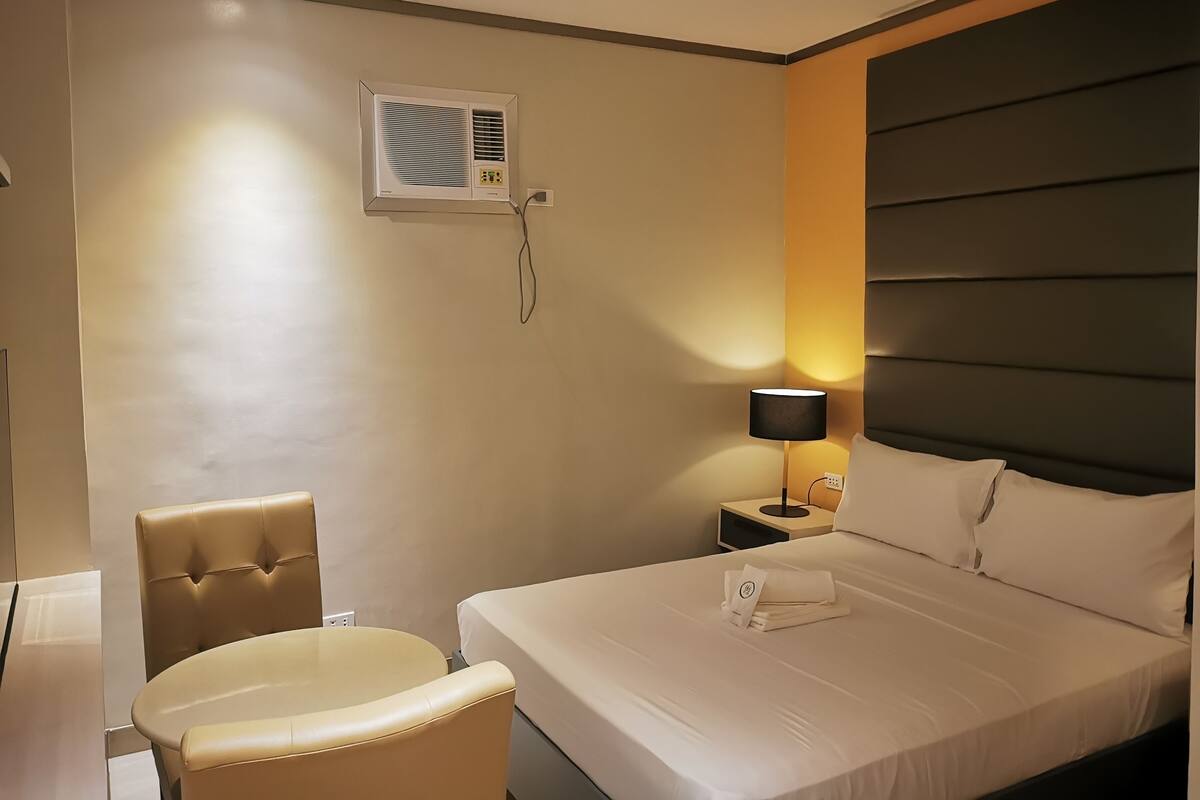 Hotellihuone, jossa on sänky, tuoli, yöpöytä, lamppu ja ilmastointilaite.