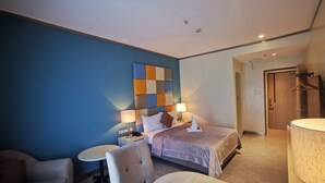 A modern hotel room with a large bed, a bedside table with a lamp, a chair, and a small round table.