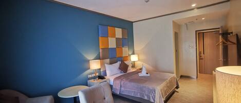 Una habitación de hotel moderna con una cama grande, una mesita de noche con lámpara, una silla y una pequeña mesa redonda.