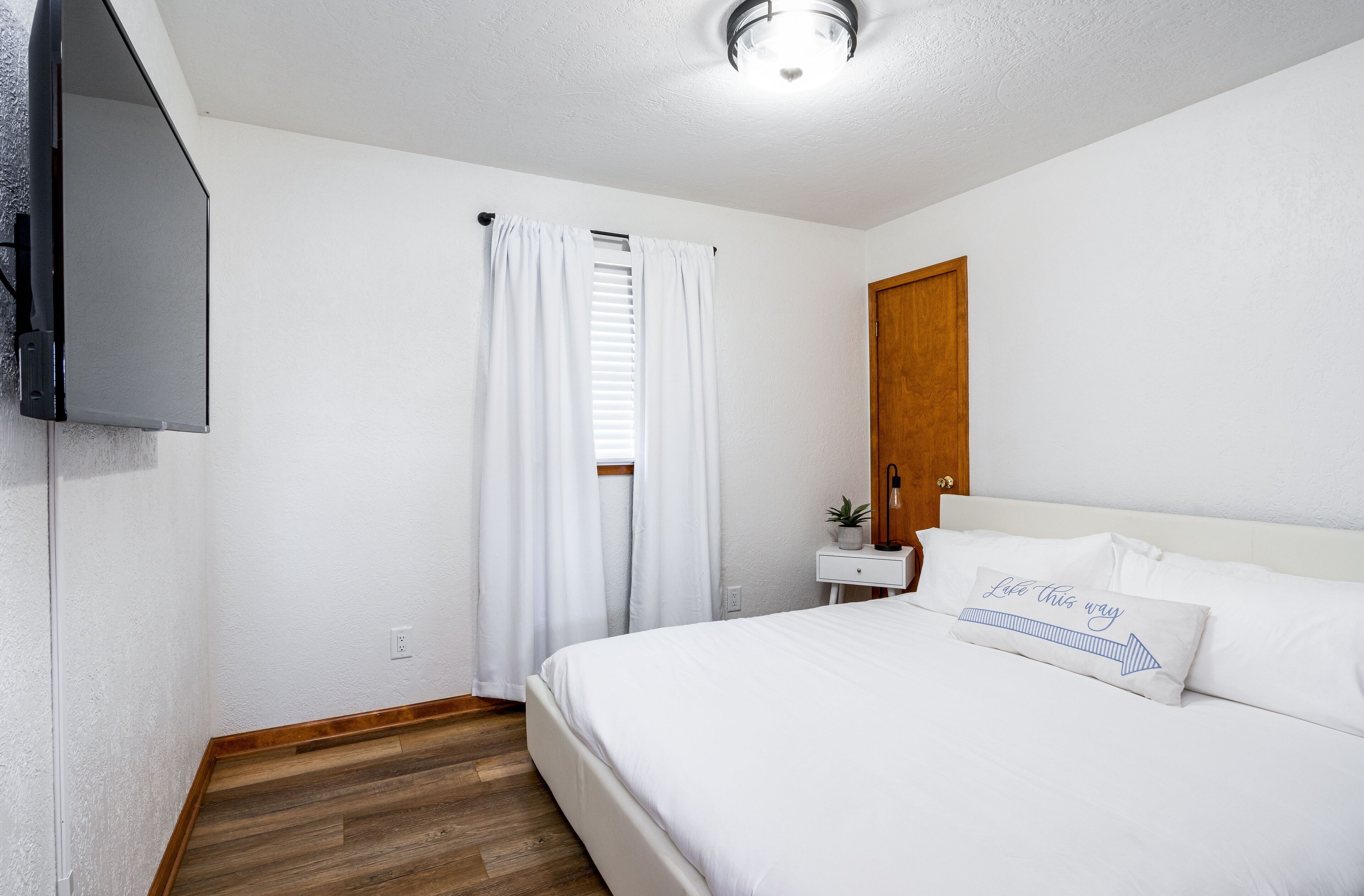 Une chambre à coucher avec un lit, des rideaux blancs, un plancher en bois, une table de chevet avec une lampe et un téléviseur fixé au mur.