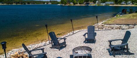 Un espace de détente aménagé au bord du lac, avec des fauteuils Adirondack et un foyer, entouré d’un chemin de gravier et offrant une vue sur le lac et les maisons situées au loin.
