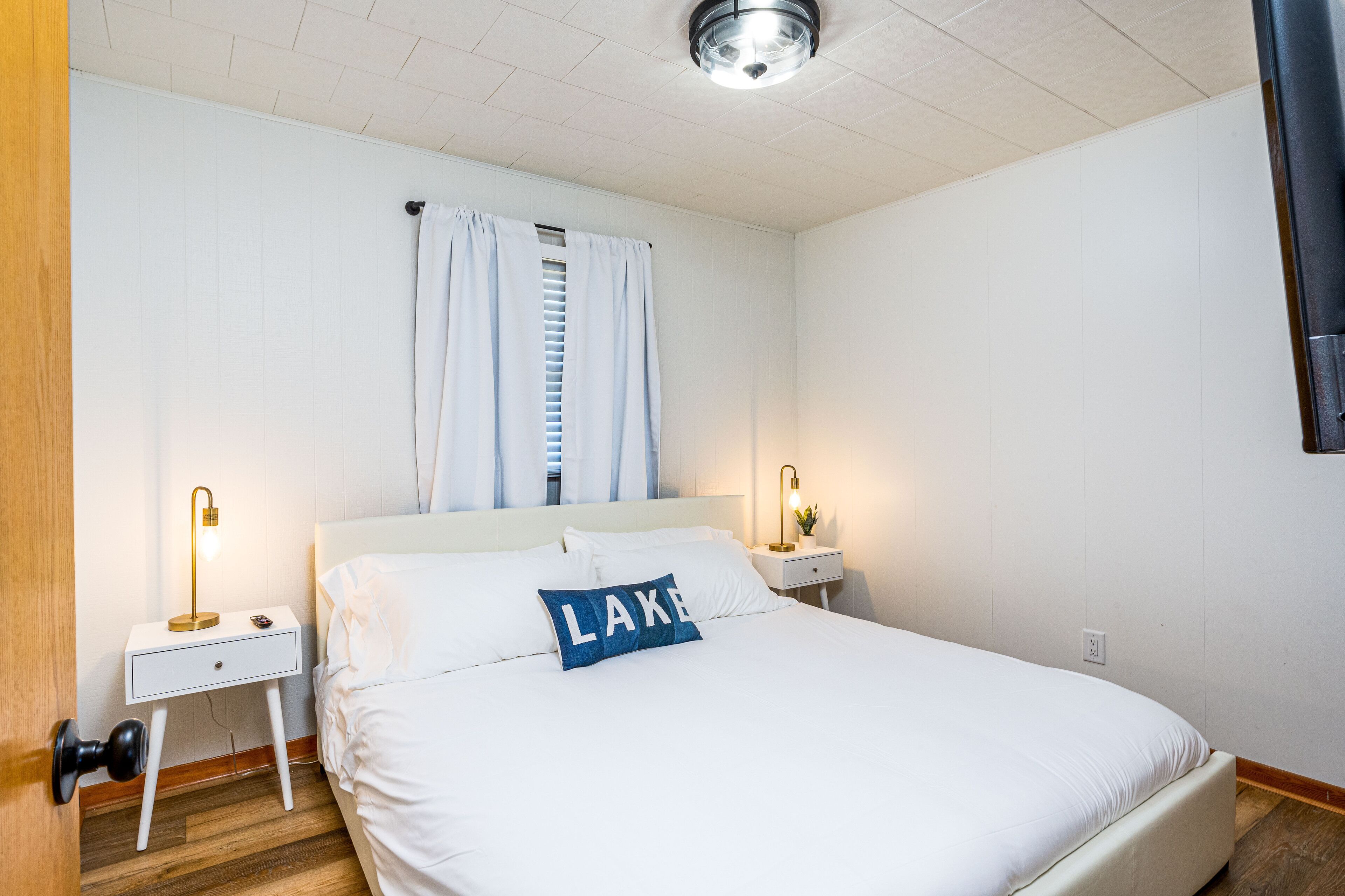 Una camera da letto con un letto, tende bianche, un comodino con una lampada e un punto luce a parete.