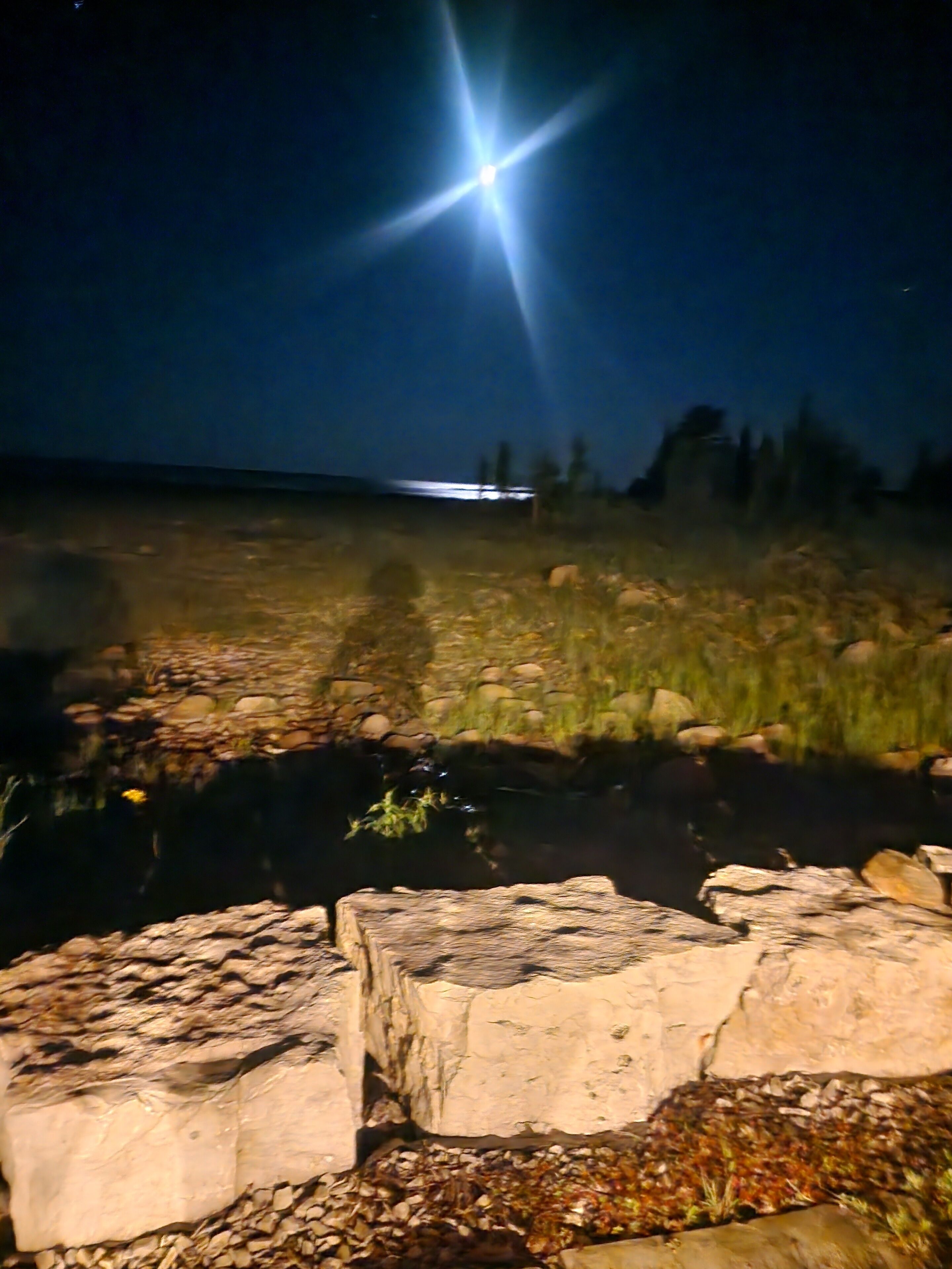 Scena notturna all'aperto con una fonte di luce intensa, un sentiero roccioso e una piccola distesa d'acqua.
