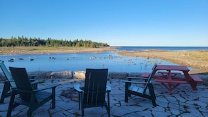 En uteplats vid sjön med Adirondack-stolar och ett picknickbord, med utsikt över en lugn sjö med ankor och en skog i bakgrunden.