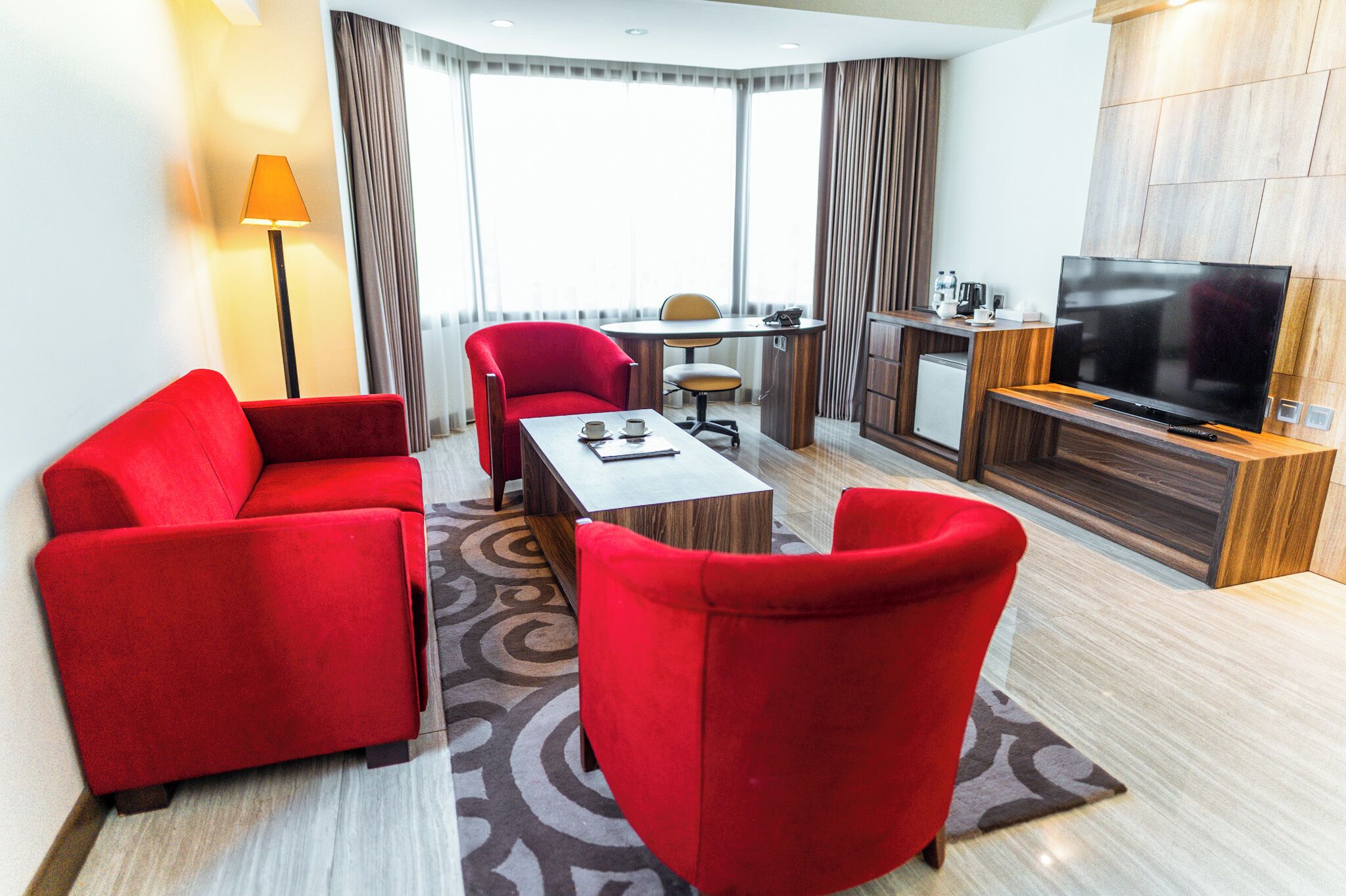 A modern hotel room with a red sofa set, a dining table with chairs, a TV, and a small kitchenette.