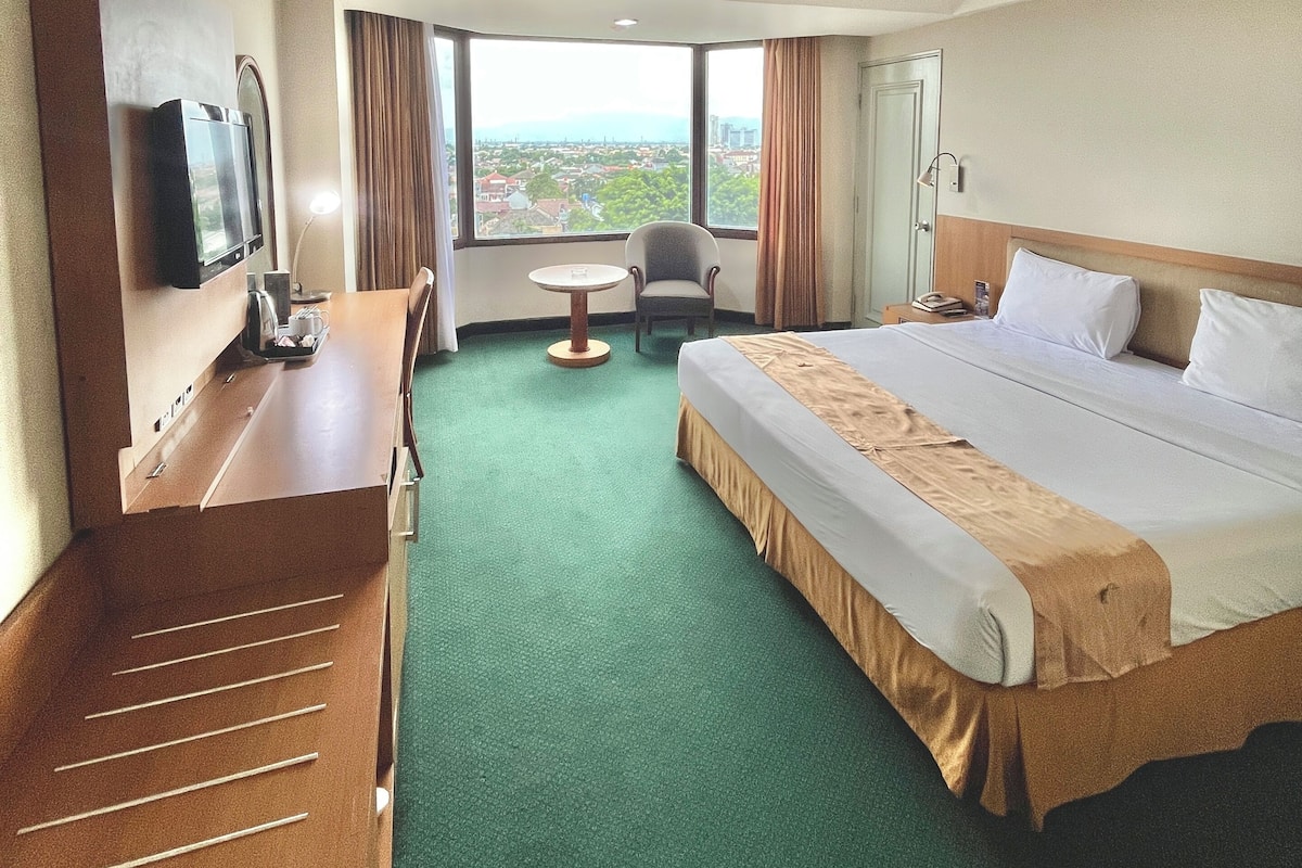 A hotel room with a large bed, a desk with a flat-screen TV, a chair, a small table, and a view of the city through large windows.