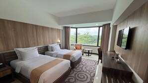 Quarto de hotel com duas camas, uma escrivaninha, uma cadeira, uma televisão e uma janela grande com vista para o verde.
